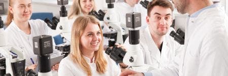 Estudiantes con batas de laboratorio aprendiendo en la sala de microscopios con varios microscopios verticales equipados con cámaras digitales. El profesor sostiene una tableta cuya pantalla muestra una imagen magnificada.