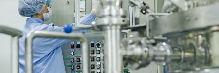 Pharmaceutical plant employee wearing personal protective equipment (PPE) checking the readouts on processing equipment with a stainless steel processing pipe in the foreground